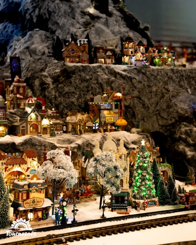Village miniature de Noël enneigé avec maisons illuminées, sapin lumineux et rails de train sur une montagne décorée